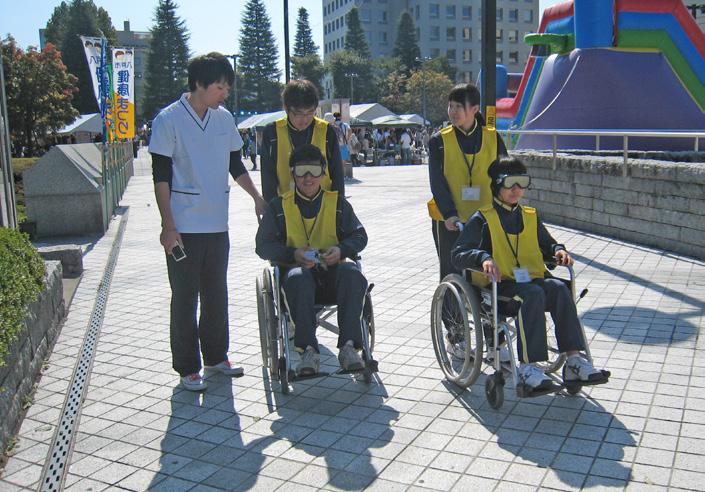 ゴーグルを装着して車いすに乗り押してもらう擬似体験をしているジャージ姿の学生の写真