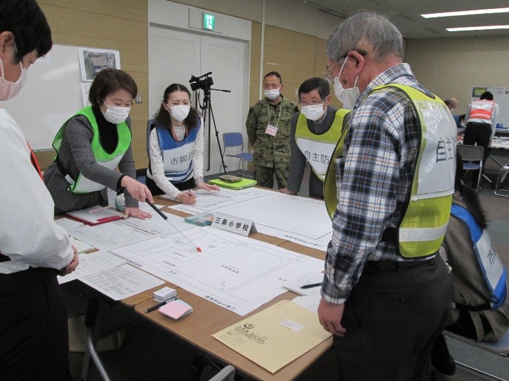 会議室でビブスを着けた参加者7名が、テーブルに広げた訓練施設の平面図を囲み、話し合っており、そのうち1名が指示棒を使って平面図の一部を指している写真