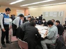 会議室で、避難所運営訓練の参加者が6名から7名ずつのグループに分かれて、学校の平面図を広げたテーブルを囲んで座り訓練している写真
