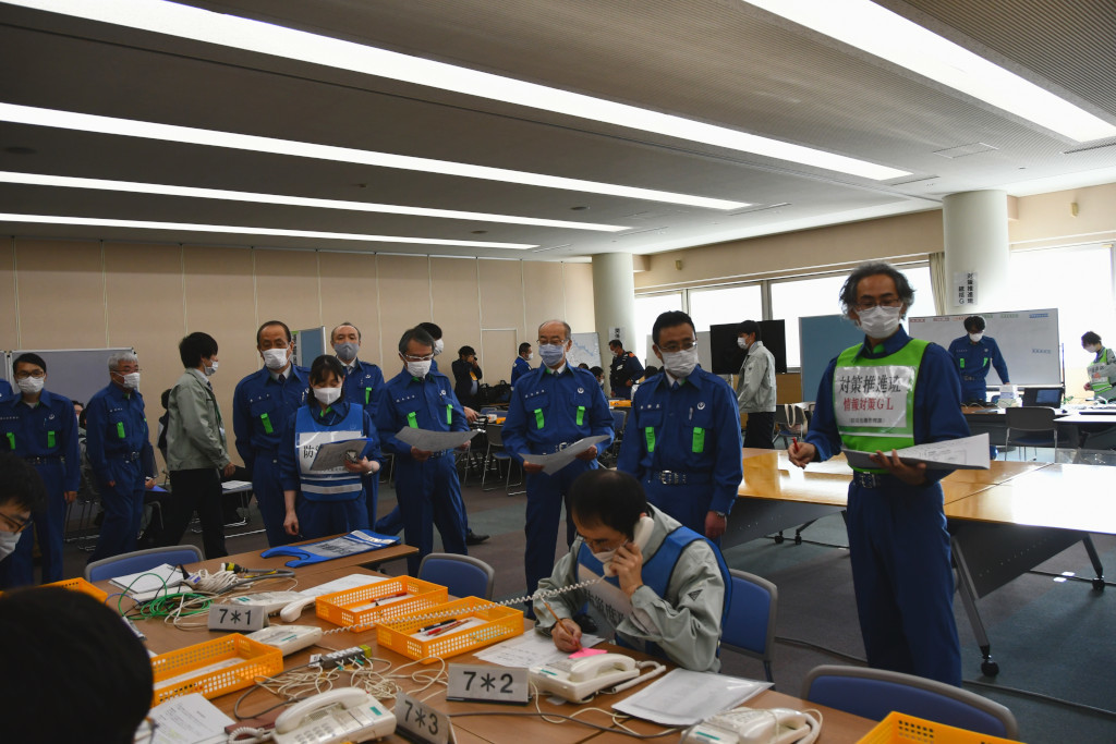 訓練会場となった会議室で防災服を着た八戸市長を含めた10名ほどが視察に来ており、ビブスを着た担当者が市長に訓練の状況を説明している写真