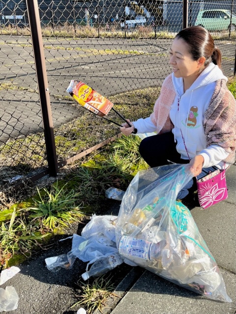 駐車場フェンスの脇でポイ捨てされた紙パック飲料の空容器をトングでつかんでいる写真