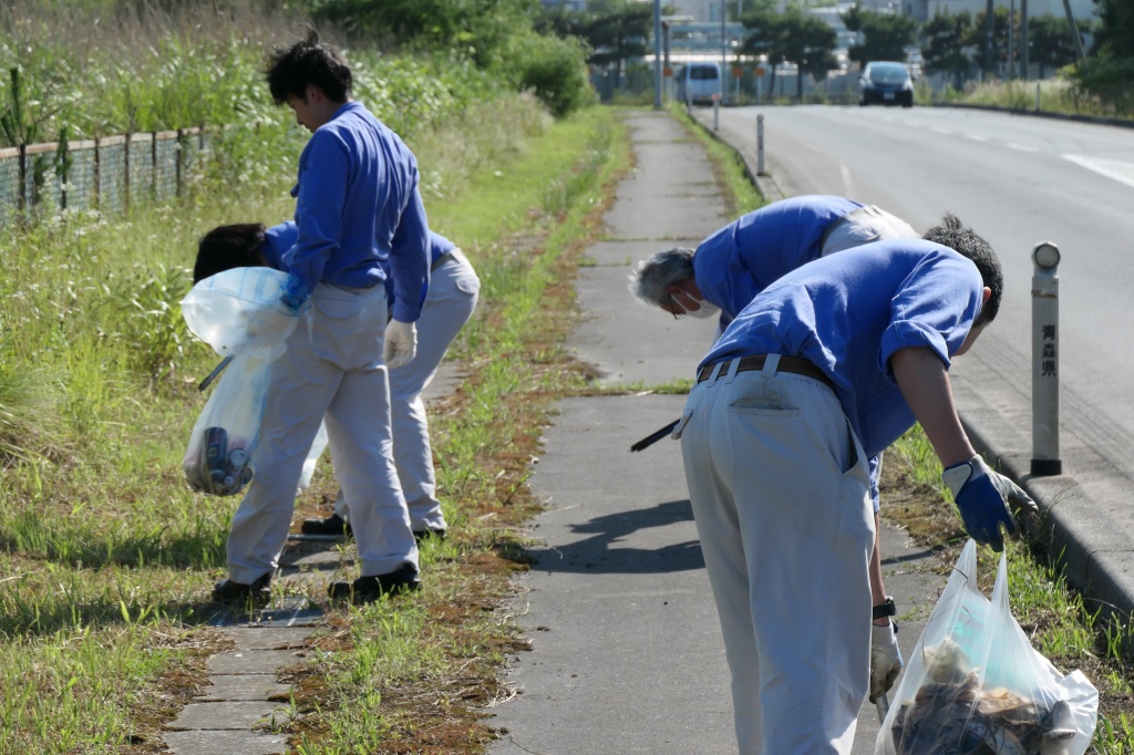 作業服姿で草の生えた歩道のごみをトングで拾う4名の写真