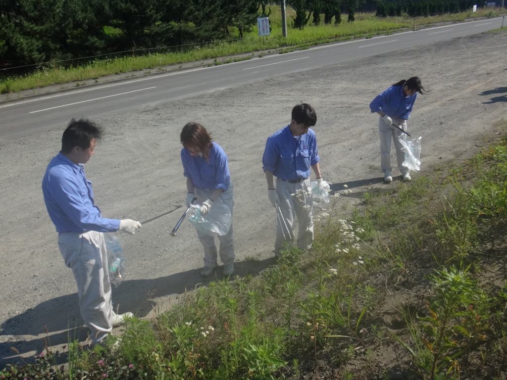 作業服姿で道路脇の草地からごみを拾う4人の写真