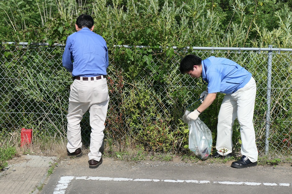 フェンスの向こう側が背の高い草木で密集している場所。フェンス下のごみを拾っている二人の写真。