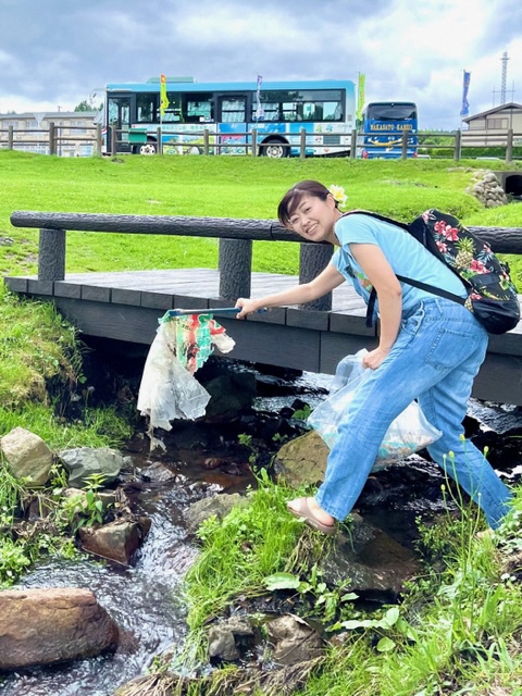 端のかかっている水の流れている場所で、トングで掴んだごみをこちらに見せている女性の写真