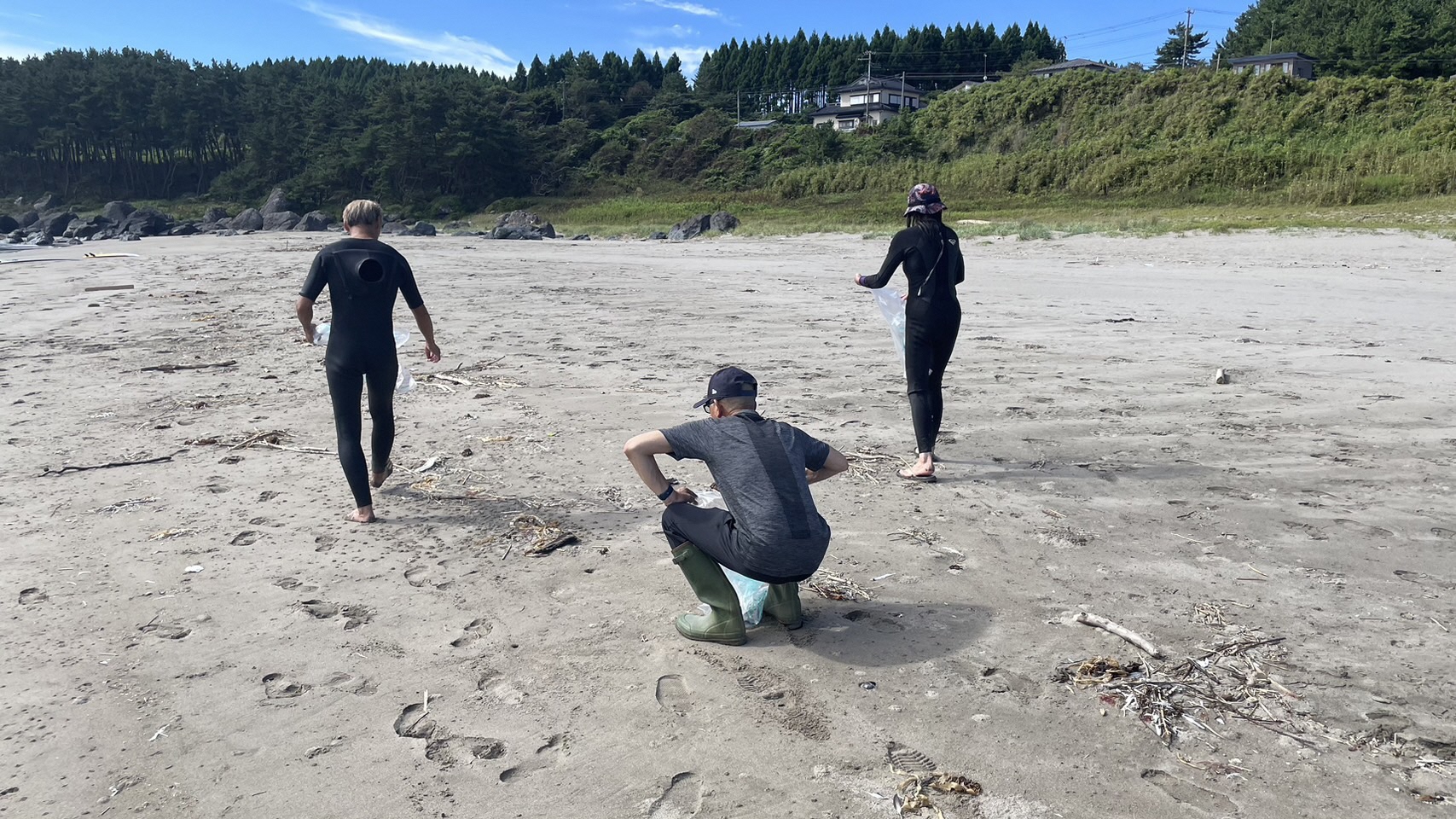 広がる砂浜に打ち上げられた枝木、ごみを拾う三人の写真