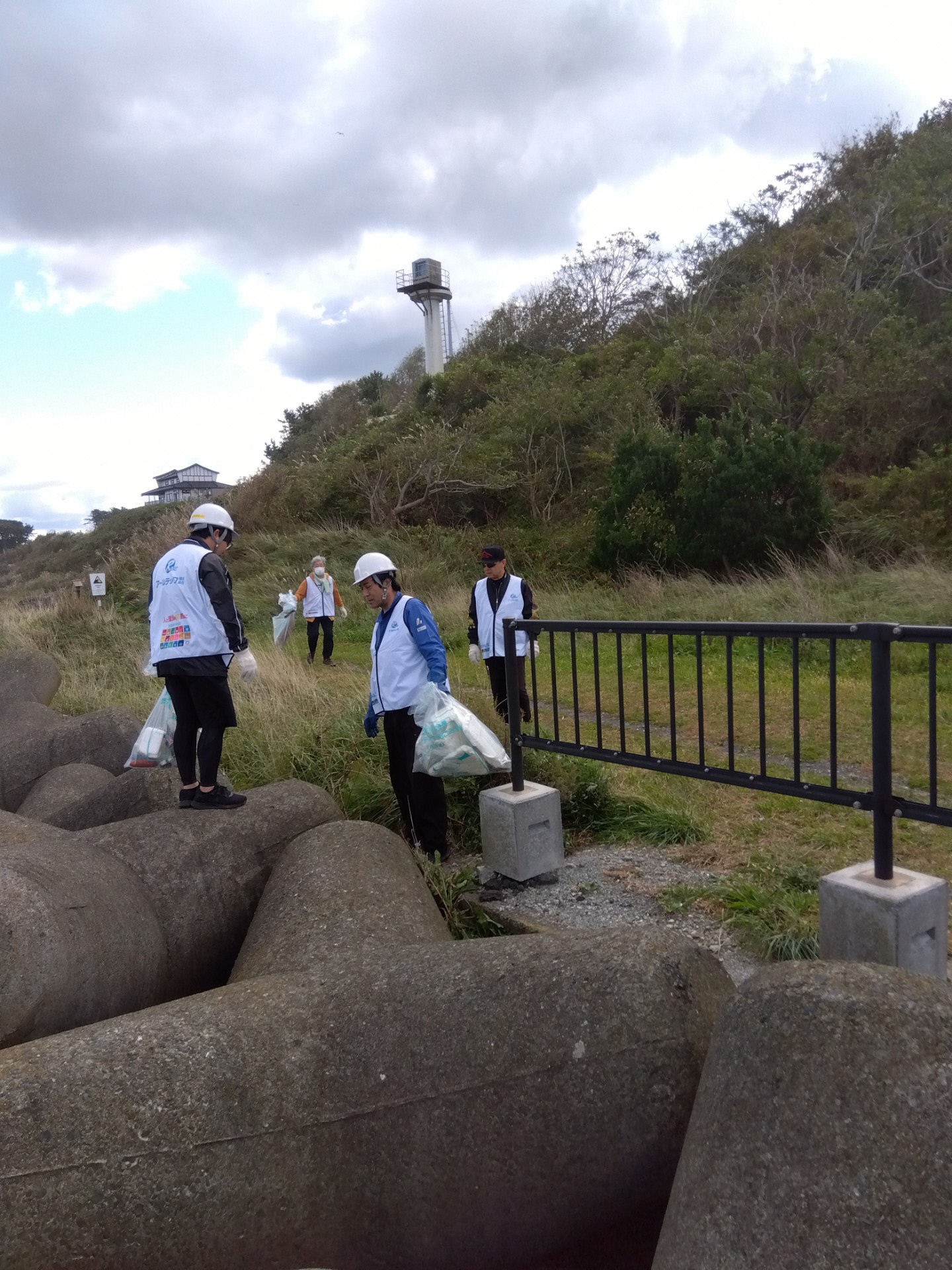 草地とテトラポットの間に立ち、ごみを拾う4人の写真