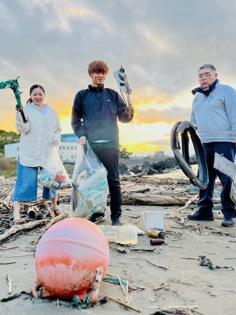 夕日を背景に砂浜で集めたごみをトングで掴んで見せている、女性、男性二人の写真