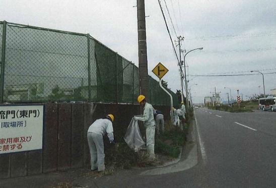 会社のフェンスの外側にある歩道のごみを拾っている作業服、ヘルメット姿の5人位の写真
