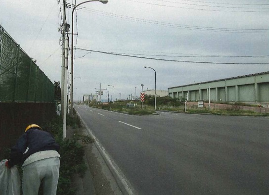 広い道路。歩道の草の生えている場所のごみを拾っている作業服、ヘルメット姿の人の写真