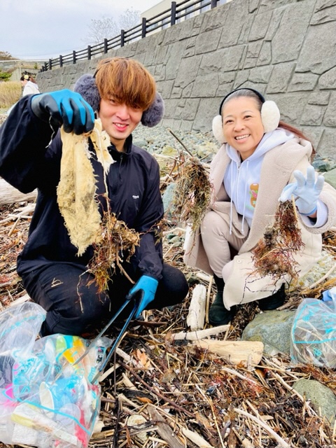 砂浜に流れ着いた木々の中から、細かいごみを分別しこちらに見せている男性、女性二人の写真
