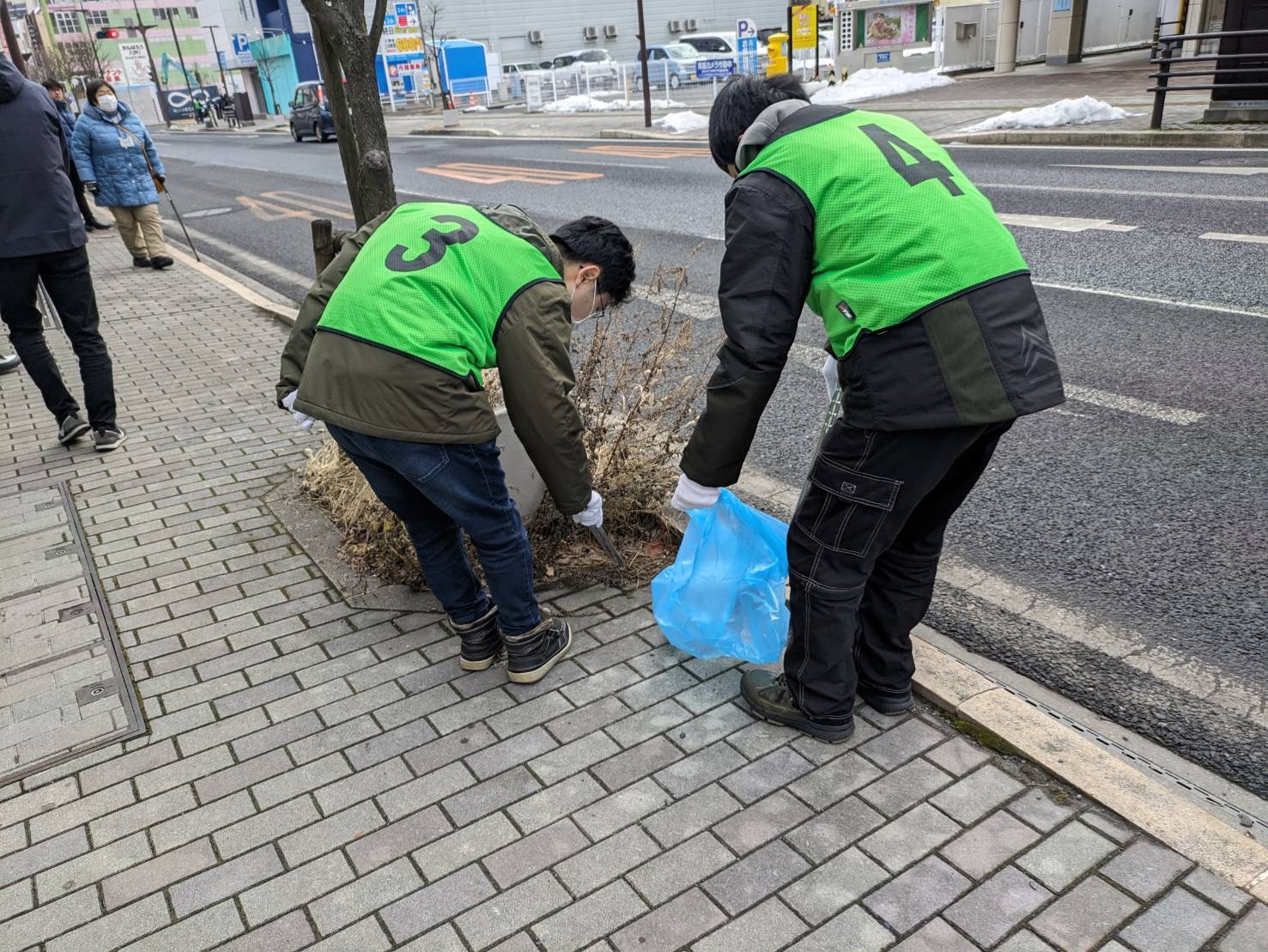 数字の書かれたビブスを着用し、歩道の植え込みの中のごみを拾っている写真