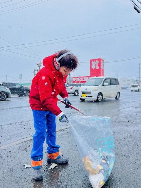 車の行きかうすぐ脇の歩道で、耳当てをしてごみを拾う男児の写真