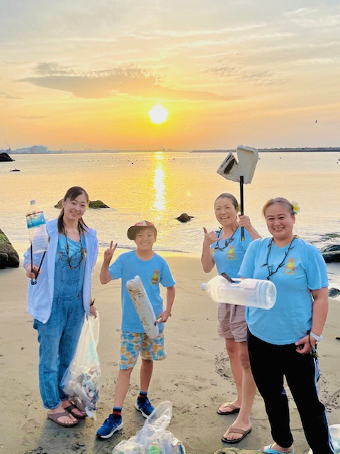 参加者4人が拾ったごみを手にしている写真。後ろは海に夕日が沈んでいく光の道ができています。