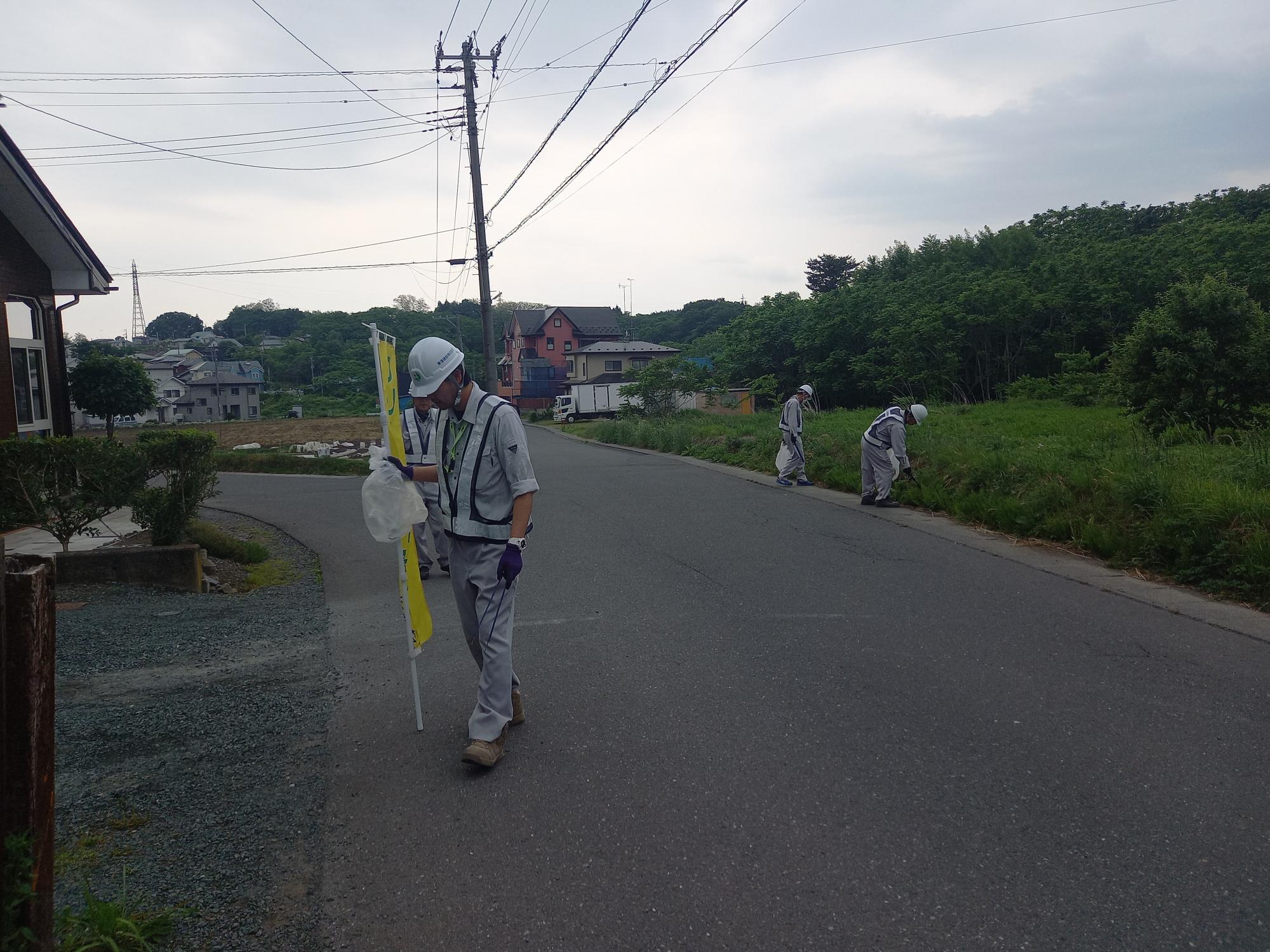 奥に緑の広がる道路でクリーンパートナーの幟を手に、活動する作業服姿の人たち