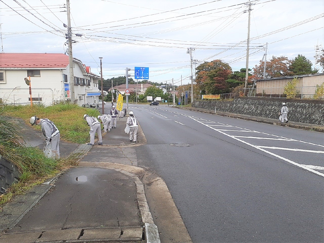 道路脇の歩道のごみを集めている作業服、ヘルメット、安全帯を着用している参加者の写真。