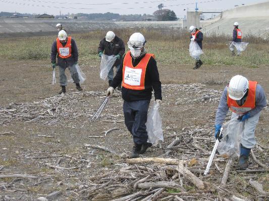 木々の漂着した川岸。ヘルメット、作業服にビブスを着用した参加者がトングを手にごみを拾っている写真。