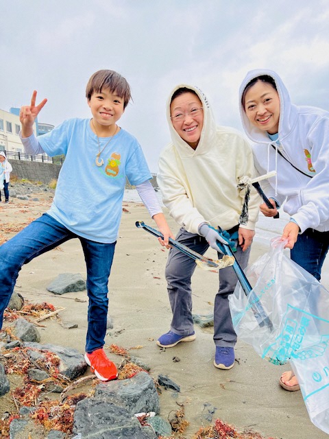 砂浜で拾ったごみをボランティア用ごみ袋に入れている、女性二人とピースサインを出している男児の写真。