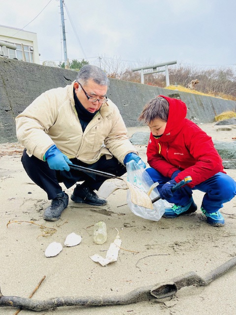 砂地にいる防寒着の男性と男児の写真。トングで拾ったごみをボランティア用ごみ袋に入れている。