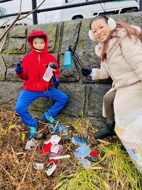 道路下の雑草の上のプラスチックごみが沢山あり、トングで拾っている耳あて