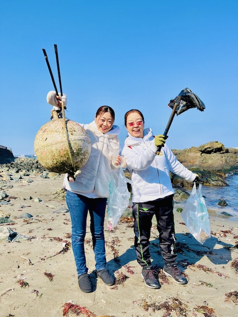 青空の砂浜。集めた浮きと長靴をトングで掲げている女性二人の写真。