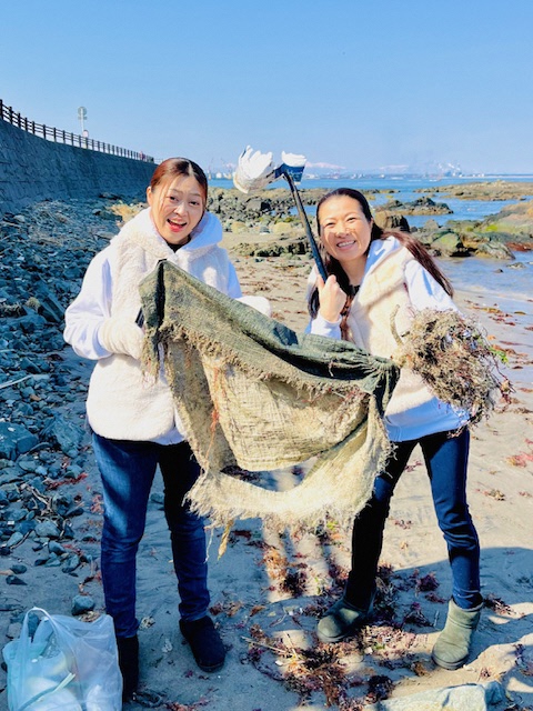 青空の海岸。集めたごみをトングで掲げている女性二人の写真。