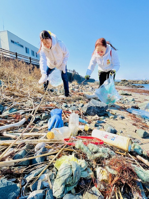 青空の海岸。たくさんの漂着ごみを拾う女性二人の写真。