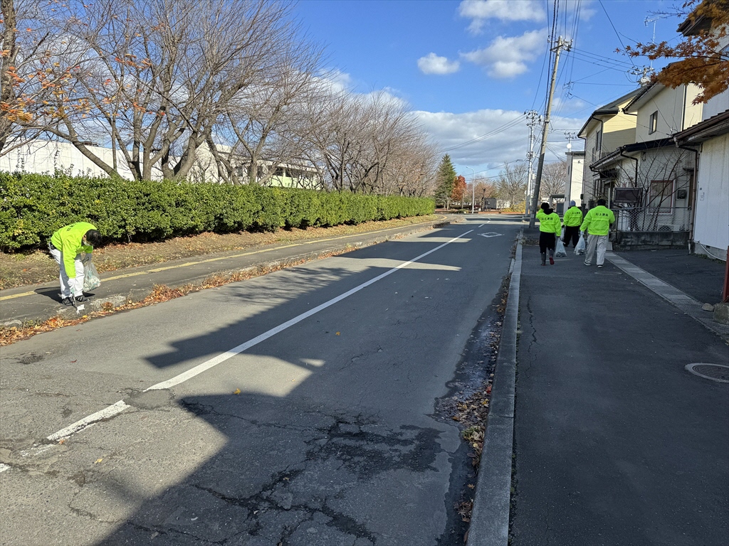 社名入りの黄緑色のジャンパーを着て、歩道のごみを拾っている写真