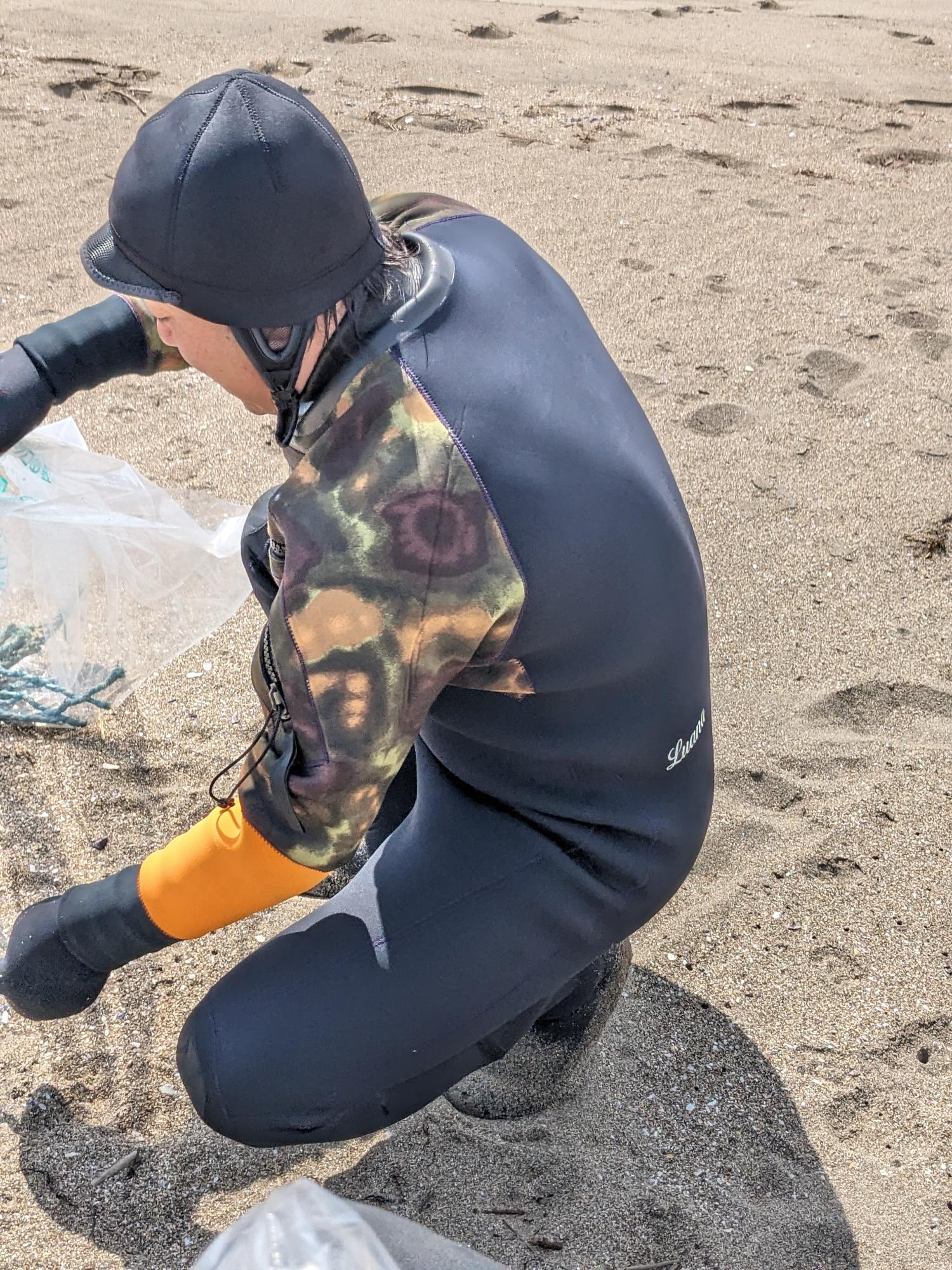 ウエットスーツを着て砂浜のごみを拾う男性の写真