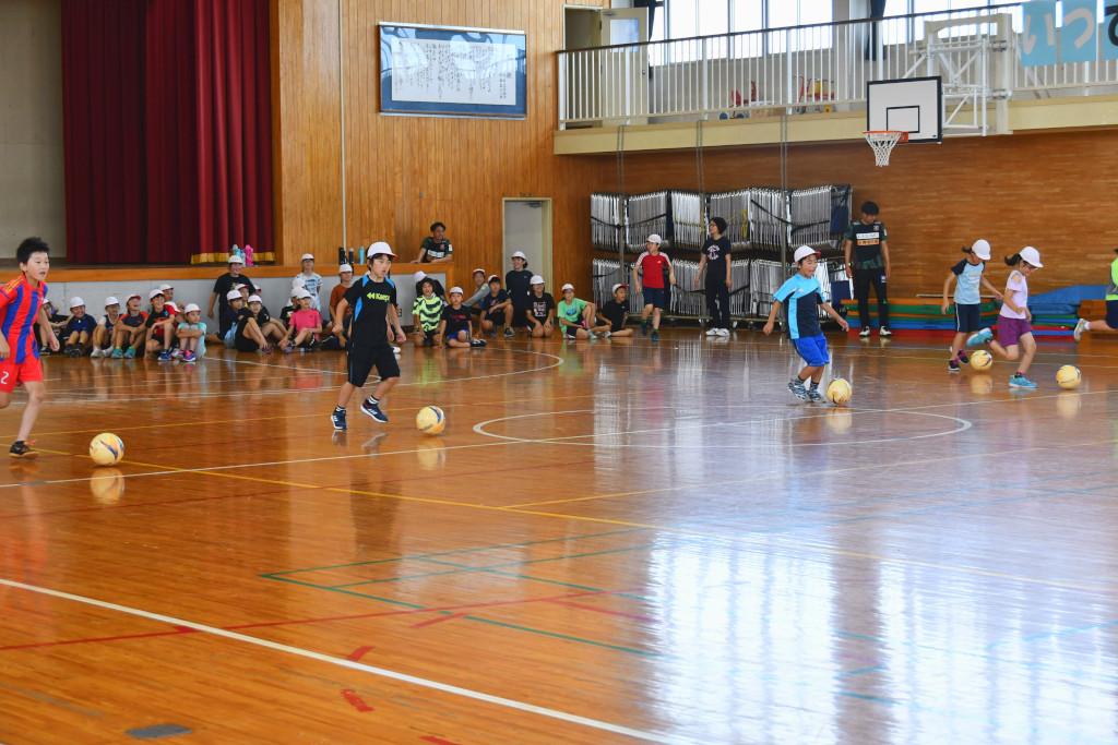 体育館にて舞台前に座っている生徒たちの前でサッカーのドリブルをしている生徒が数名写っている写真