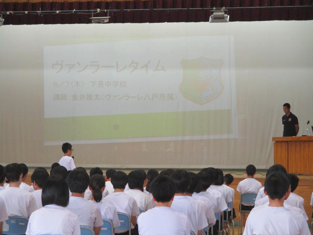 体育館の舞台に向かって椅子に座っている生徒たち、うち1名が立ってマイクをもって舞台に立っている男性に御礼の挨拶をしている写真