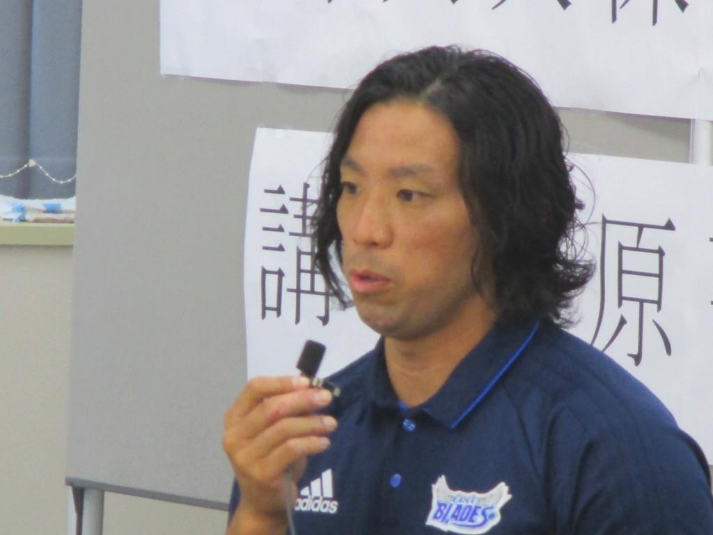 室内にてピンマイクをもって座って話をしている男性の写真