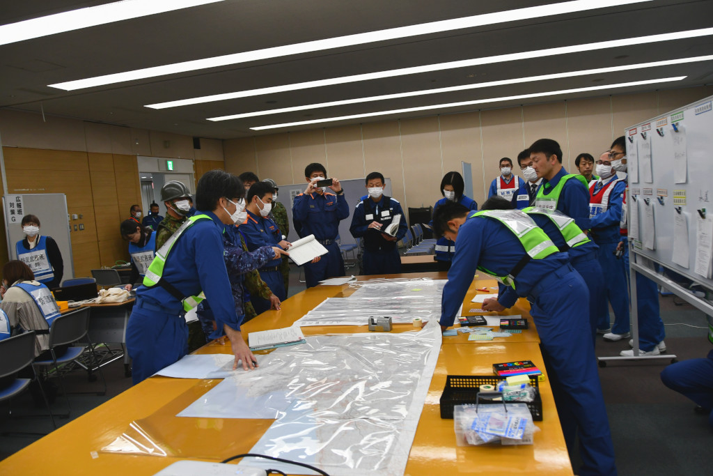 会議室の中央に地図を広げたテーブルと、その右側にホワイトボードが設置され、ビブスを着た市職員と自衛隊、消防、警察の担当者が地図を囲んで話し合っている写真