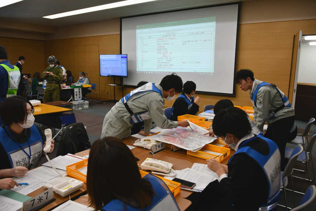会議室の中央にはスクリーンが設置され、その右隣にはモニターが設置され、被害状況や避難所の避難者状況が表示されており、その周辺でビブスを着た参加者がテーブルを囲んで作業をしている写真