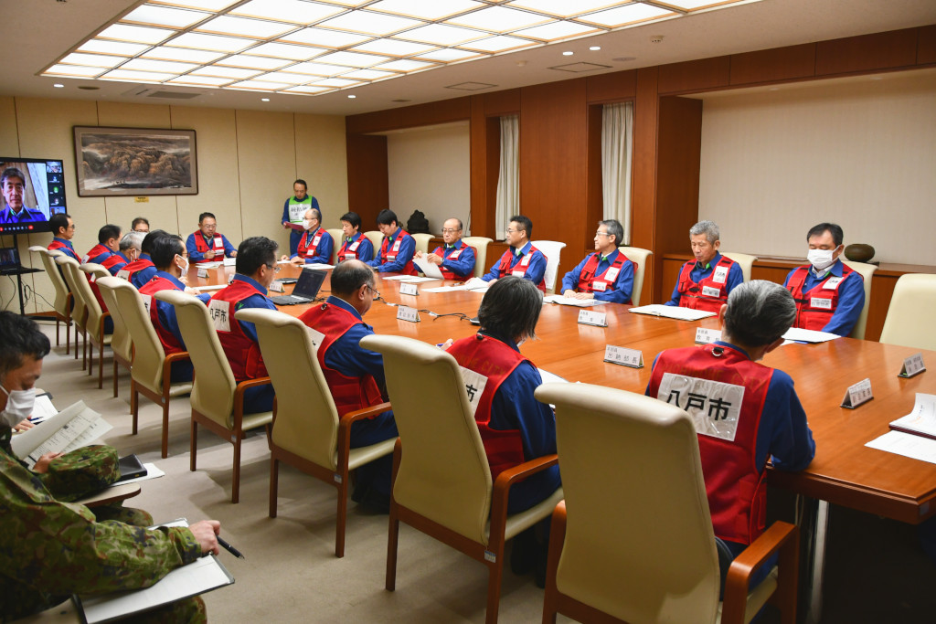 八戸市庁議室で、災害対策本部員20名ほどが赤いビブスを着て、会議用テーブルを囲んで座り、テーブル奥に司会進行役が立って会議を進めている写真