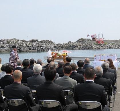 大久喜漁港で神主さんが前に立ち鳥居再建式が執り行われている写真
