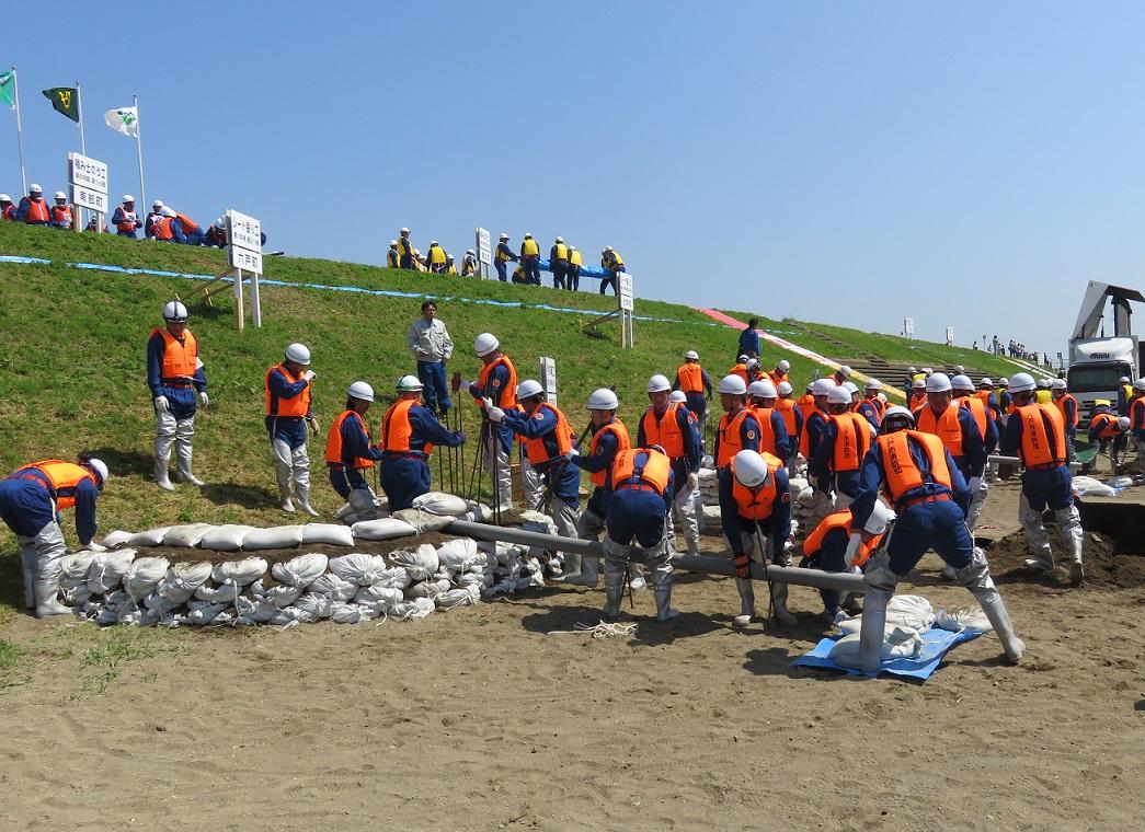 河川敷でオレンジの防災チョッキを着た参加者達が土嚢を積んで水防演習をしている写真