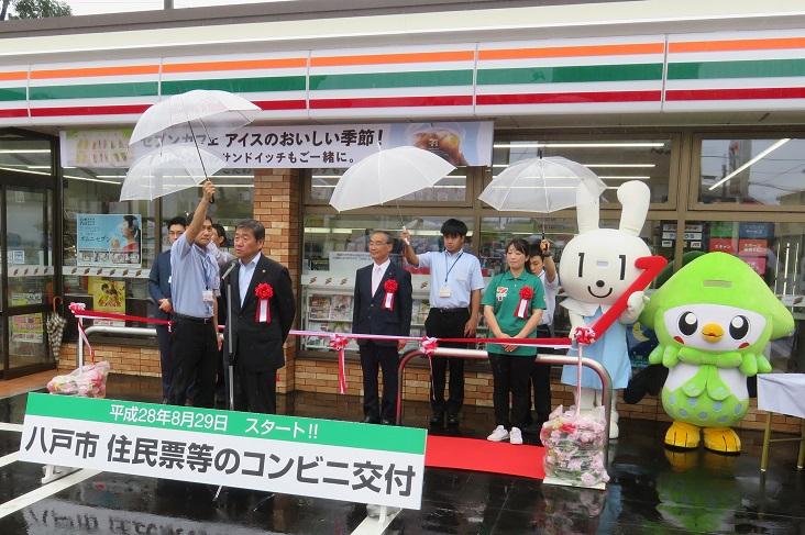 「コンビニ交付セレモニー」で雨の中傘を差してもらいながらコンビニの前で挨拶をしている市長の写真