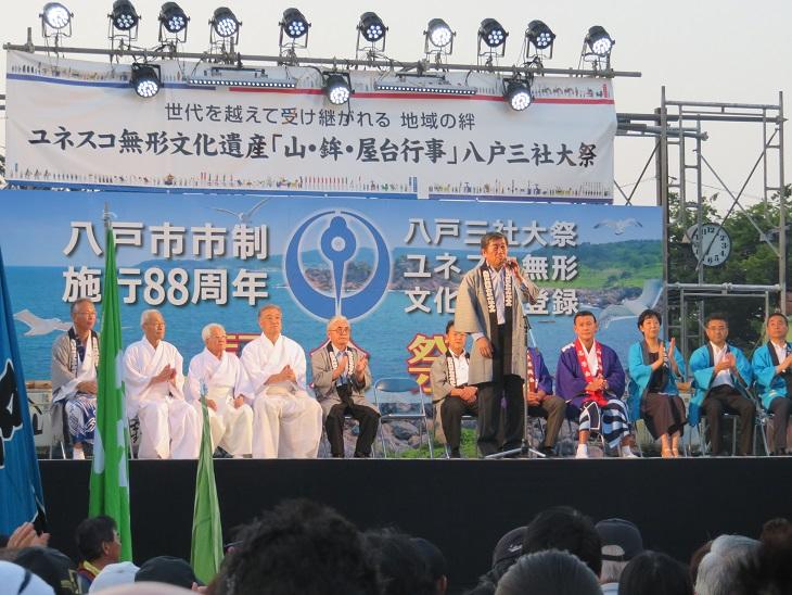 ユネスコ無形文化遺産登録記念祭で舞台上に10名ほどの袴や法被を着た人々が座っていおりその前で挨拶をしている市長の写真