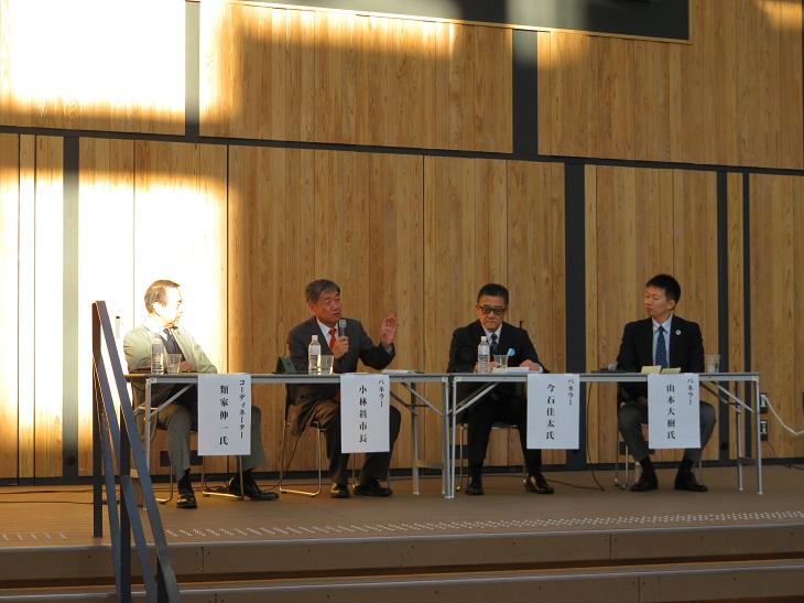 防災フォーラムでゲスト3人と長机に座りパネラーとしてマイクを右手に持ち話をしている市長の写真