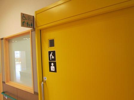 トイレの入り口左側に男女共同、車いす利用者、障がい者、赤ちゃんが一緒でも利用可能な絵文字表示のある黄色のスライドドアの多目的トイレの写真