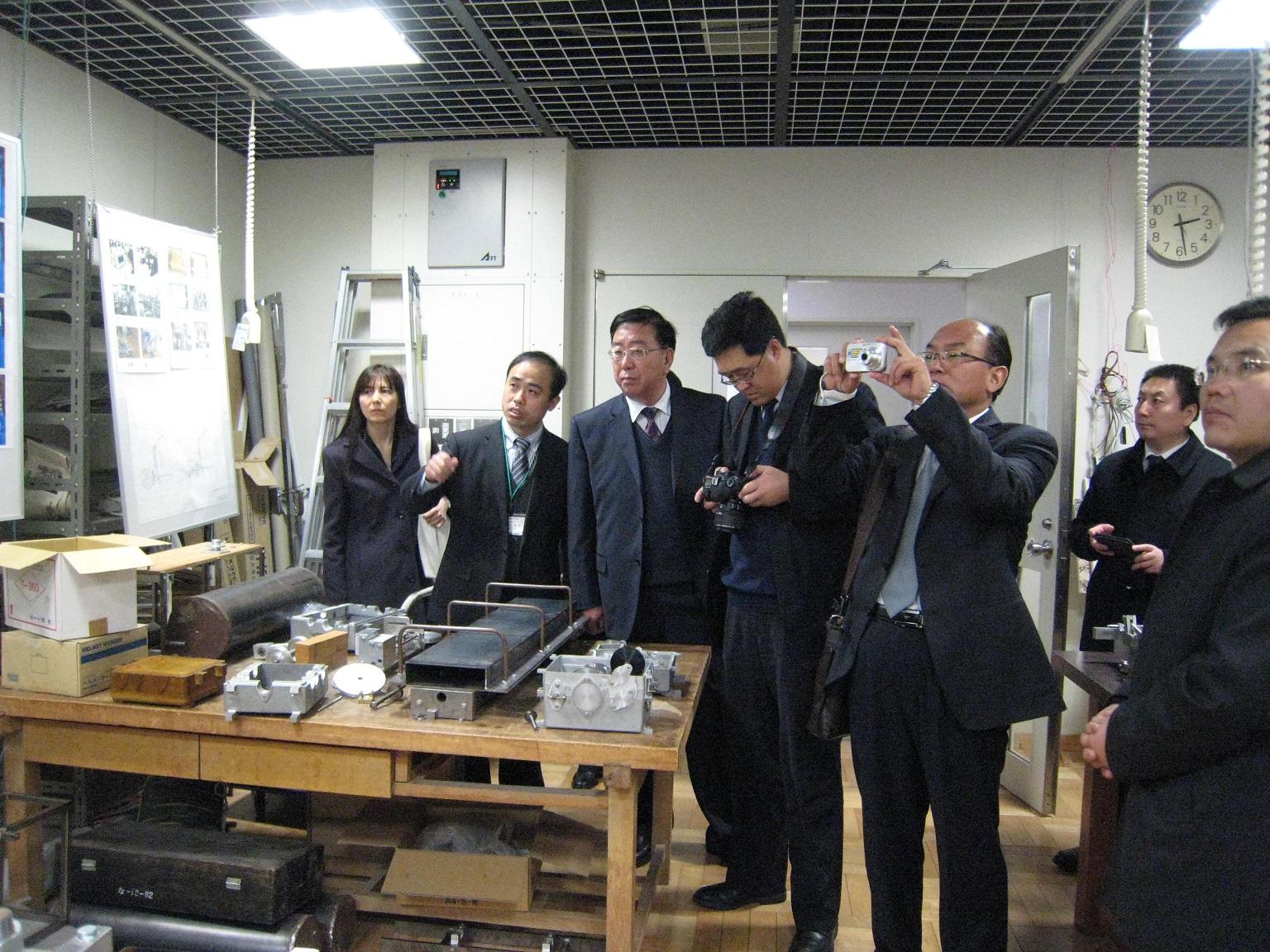 7人のスーツの男女が専門学校のさまざまな工具が置かれた部屋で写真を撮っている写真
