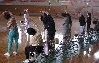 体育館で7人の参加者が講師の動きに合わせてストレッチをしている写真
