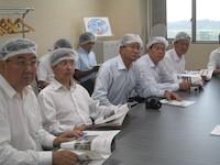 8人の男性が白い頭巾とシャツをきて机に座り話を聞いている写真