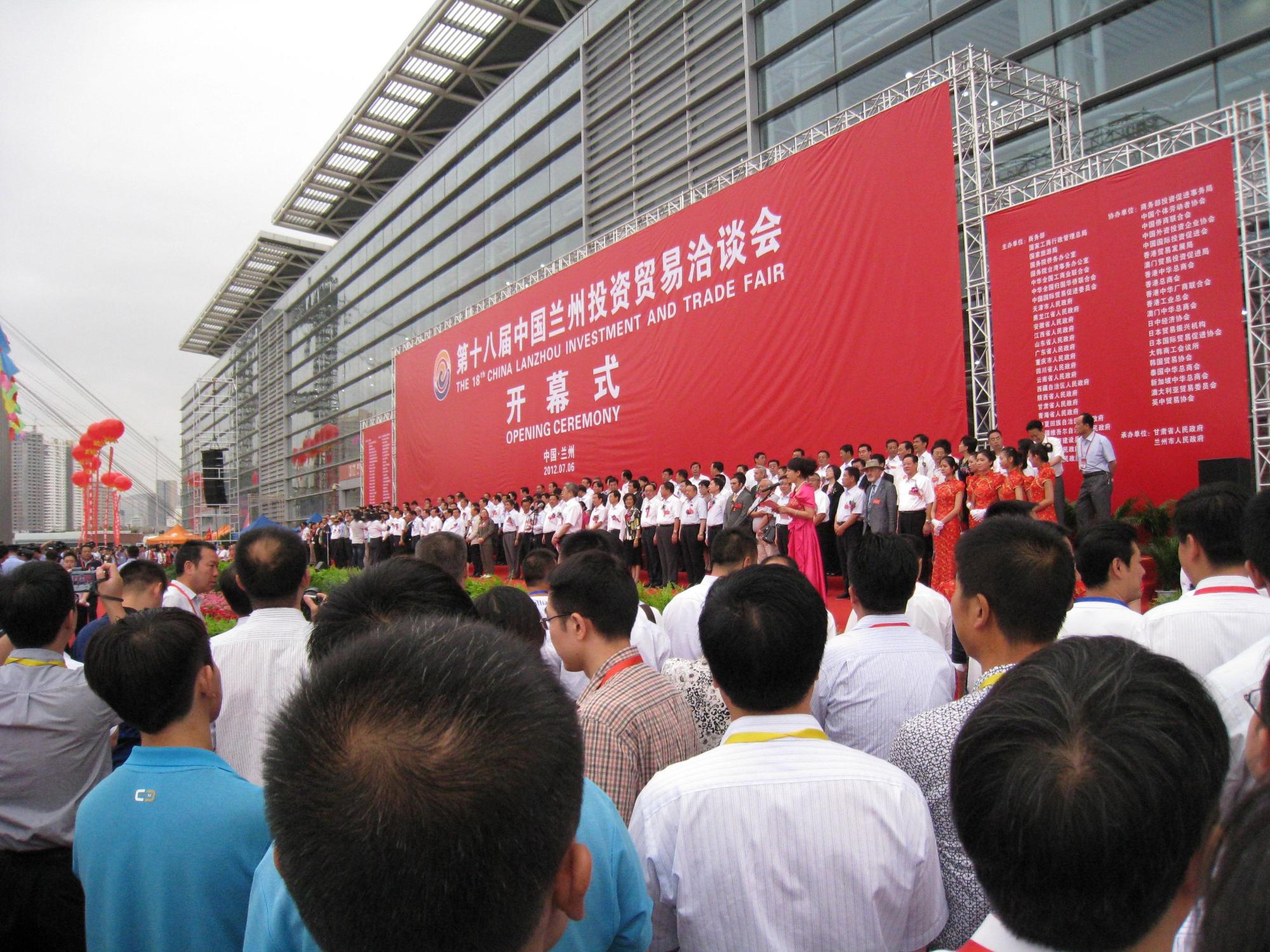 50メートルほど横幅のある朱色の横断幕の前に同じ幅のステージがあり、その壇上に百人を超す白いシャツを着た出席者が立っている写真