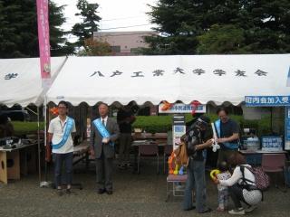 八戸工業大学学友会の白いテント前の左側でタスキをかけた男性が立っている、右に3名の大人が写っている写真