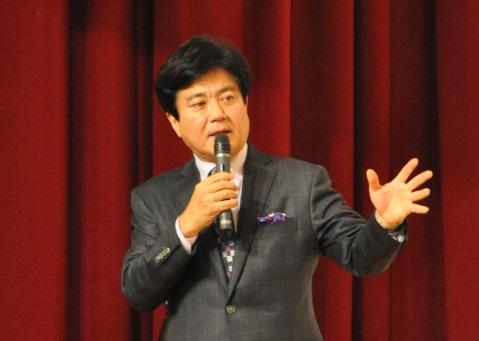 臙脂色のステージの幕の前で開いた左手を聴講者に向けてスピーチをするグレーのスーツを着た男性の写真