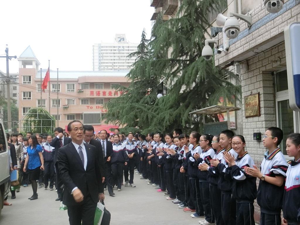 学校を訪問している複数のスーツの男性が拍手で迎える生徒の前を歩いている写真
