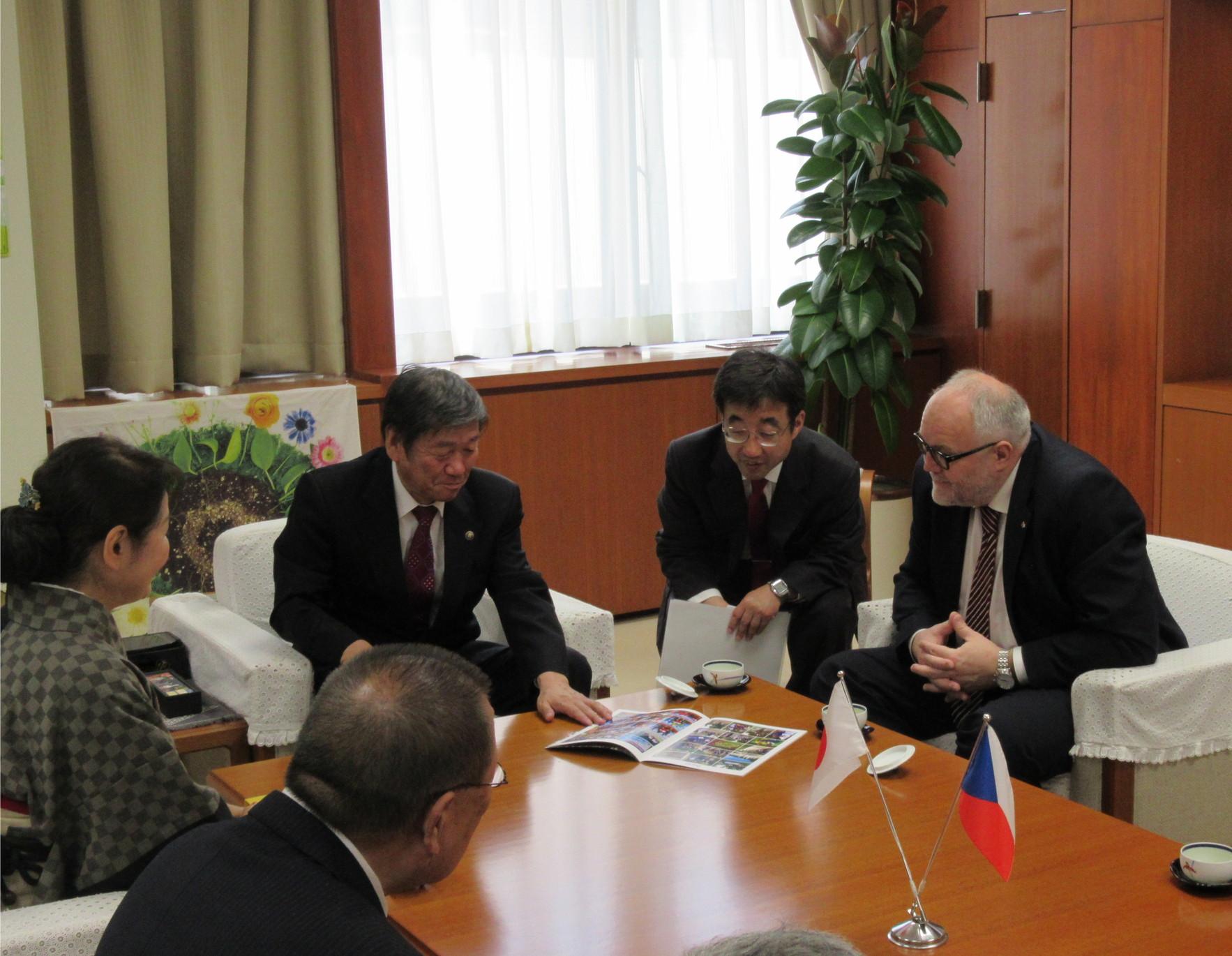 駐日チェコ共和国大使館臨時代理大使一行と小林市長が白いソファーに座って、テーブルの上のパンフレットを見て話している写真