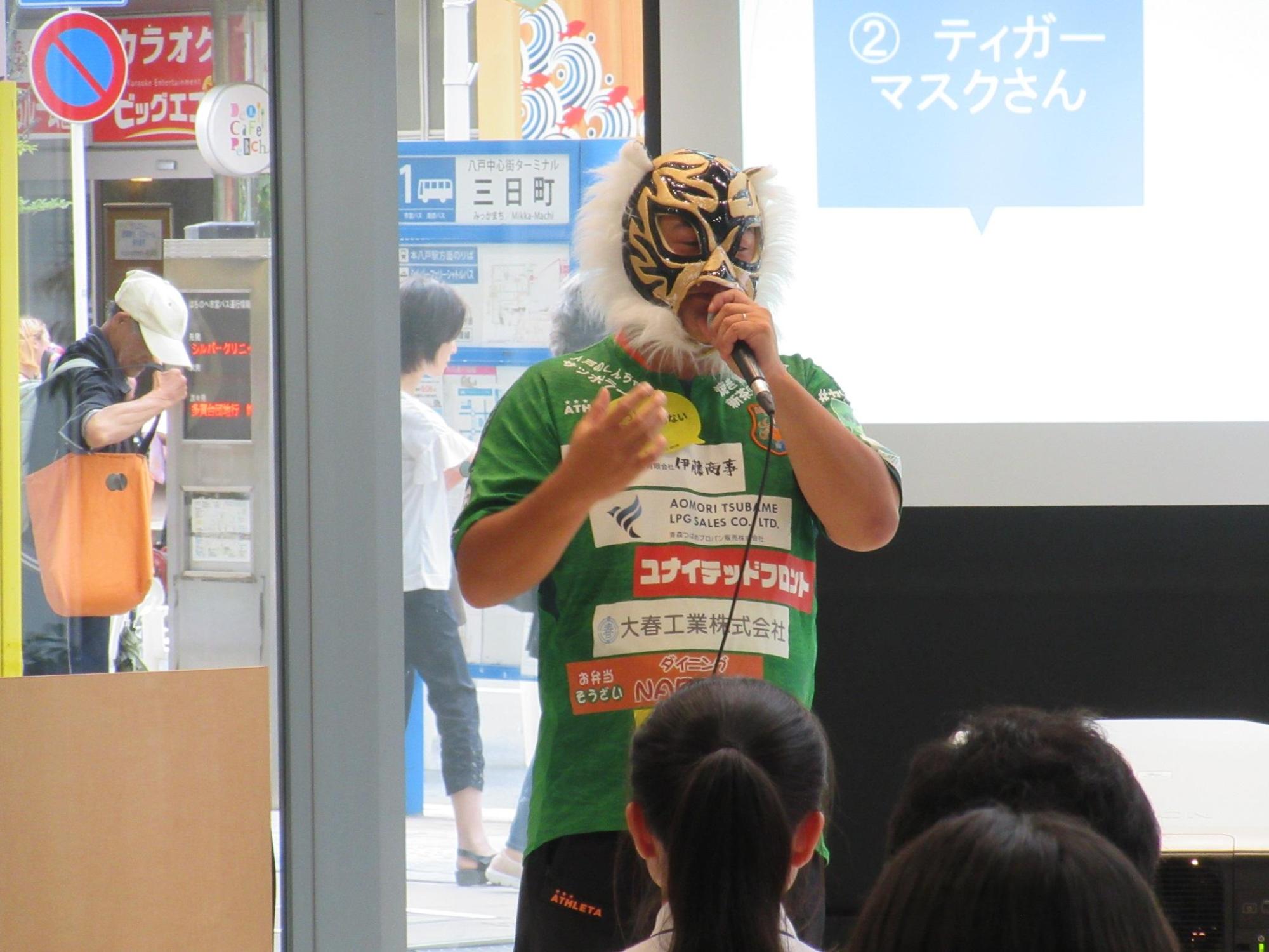 緑の服を着たマスクをかぶった男性がマイクをもって立って話をしている写真